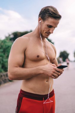 Seksi gömleksiz kaslı beyaz vücutçu motivasyon müzik listesi için hücresel teknoloji kullanıyor ve sabah antrenmanı sırasında kulak misafiri oluyor. Mobil uygulama kullanan kulaklıklı atletik adam.