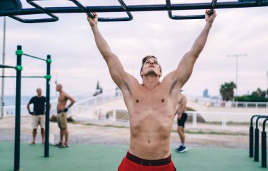 Sabah egzersizi sırasında kaslarını çalıştırmak için barfiks çeken güçlü bir erkek vücut geliştirici. Mükemmel vücut yapısı olan beyaz gömlekli bir adam şehirdeki spor sahasında enerjik bir ısınma yaşıyor.