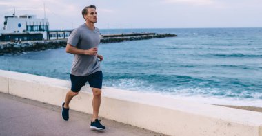 Aktif giyimde ciddi bir beyaz erkek koşucu sabah kıyı şeridinde kardiyo eğitimi alıyor sağlıklı yaşam tarzının tadını çıkarıyor kararlı erkek koşucu formda ve fiziksel olarak antrenman yapıyor.