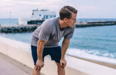 Aktif kıyafetli dalgın beyaz erkek koşucu. Kardiyo antrenmanından sonra nefes alıp veriyor. Limandaki suya bakıyor. Genç zayıf sporcu, koşudan sonra ferahlatıcı ve ferahlatıcı.