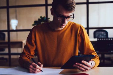 Yaratıcı erkek öğrenci, üniversite projesi için dijital tablet kullanırken sınıfta eskizlerle çalışan beyaz tenli hippi çocuk aracılığıyla araştırma yaparken ders için zaman harcıyor.