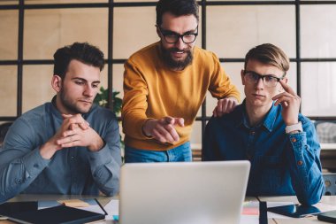 Genç erkek portresi, arkadaşlarının yanında oturmuş dizüstü bilgisayarda webinar seyrederken kameraya bakıyor. Bir grup adam, bilgisayar üzerinden araştırma yapmaya odaklanmış.