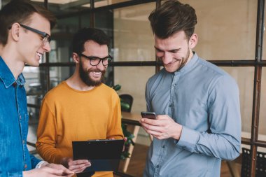 Başarılı erkek partnerler, sosyal ağda akıllı telefon cihazıyla komik fıkralar okumaktan zevk alıyor ve gülüyor, genç pozitif erkekler modern ofiste iletişim kuruyor ve teknolojiyi kullanıyorlar.