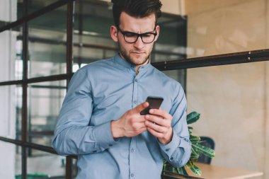 Kafası karışmış Kafkasyalı bir adam akıllı telefon cihazıyla mesaj okuyor. İnterneti tarıyor. Gözlüklü kafası karışık bir hipster modern cep telefonuyla wifi 'ye bağlı online video izliyor.