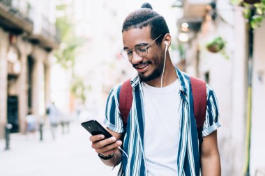 Gülümseyen Afro-Amerikan modaya uygun giyinen hippi çocuk açık havada müzik dinlemek için akıllı telefon ve ses uygulaması kullanıyor, pozitif esmer tenli adam sokakta müzik dinliyor.