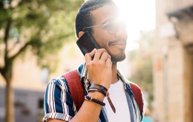 Arka taraftaki modern erkek hippi gündelik giysiler giyip şehrin orta yerinde eski binanın yakınında cep telefonuyla konuşurken tabletle dikiliyor.