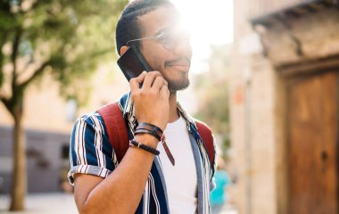 Arka taraftaki modern erkek hippi gündelik giysiler giyip şehrin orta yerinde eski binanın yakınında cep telefonuyla konuşurken tabletle dikiliyor.