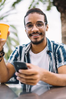 Neşeli genç etnik adam günlük giysiler ve gözlüklerle tek kullanımlık kahve içiyor ve açık kafede dinlenirken cep telefonunu karıştırıyor.