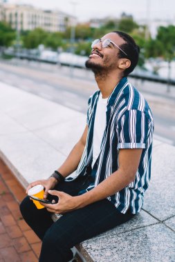 Mutlu Afrikalı genç Amerikalı hippi erkek, günlük kıyafet ve gözlüklü, akıllı telefon ve kahve bardakları var. Yaz günü şehir caddesinde taş basamaklarda otururken yukarı bakıyor.