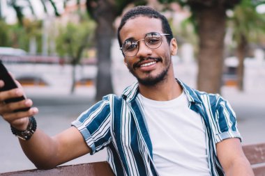 Sıradan giyinen neşeli yakışıklı hippi, kenar mahallede oturmuş içecekle dinleniyor ve akıllı telefon kullanıyor. Afro-Amerikalı bir adam kameraya bakıyor ve 4G ile mesaj ve e-posta yolluyor.