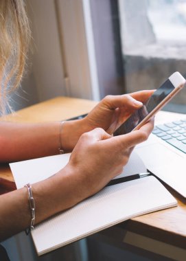 Günlük kıyafetleriyle masada oturan ve bilgisayar başında çalışırken akıllı telefon kullanan ve kafede bir fincan kahve içen bir kadın.