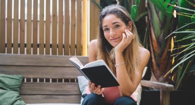 Mutlu kadın, günlük kıyafetlerini giymiş, ahşap bankta oturuyor ve hafta sonu güneşli terasta dinlenirken ilginç kitaplar okuyor.