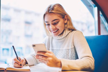 Neşeli kadın blogcu, yorumları okumak için cep telefonunu elinde tutarak yayıncılık için beğeni toplamaktan memnun, akıllı telefondan posta kontrol etmekten mutlu, test için iyi not alan hippi kız.