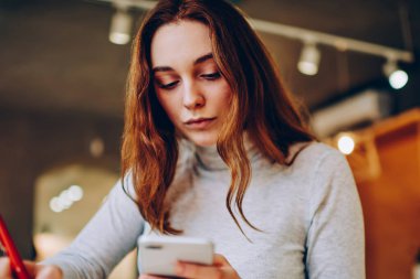 Not defterinde akıllı telefondan gelen bilgileri kaydeden konsantre bir kadın kafenin içinde uzaktan çalışan, ciddi bir kadın cep telefonu uygulamasını kullanarak not defterinde işlem yapıyor.
