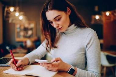 Ciddi kadın serbest çalışan not defterinde cep telefonuyla iletişim kuran, birlikte çalışma alanında çalışan, teknolojiyi ve bilgi aramak için kablosuz bağlantıyı kullanan genç bir kadın.