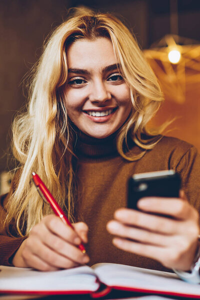 Портрет жизнерадостной хипстерши, пишущей в блокноте и держащей смартфон для чата, успешной блогерки, редактирующей текст статьи в блокноте перед публикацией в социальных сетях по телефону