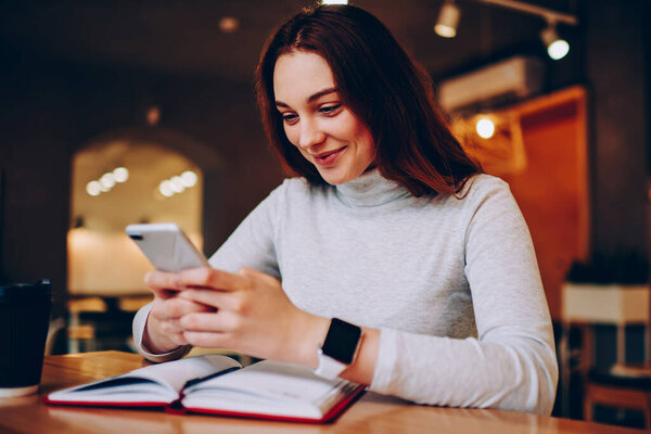 Позитивная девочка-подросток отправляет смс-сообщение бойфренду, проводящему время в интерьере кафе для обучения, веселая молодая женщина, взволнованная получением скидки на использование 4G Интернета в сетях сотовой связи