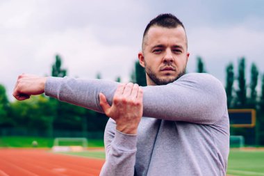Gerçekten formda sakallı genç bir antrenör kollarını esnetiyor. Stadyumda hava bulutluyken sıkı çalıştıktan sonra dinleniyor.