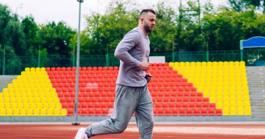 Aktif koşan genç yakışıklı erkek koşucu formda kalmak için sabah antrenmanında stadyumda koşuyor, eşofmanlı kararlı güçlü bir adam dışarıda yağ ve kalori yakmak için antrenman yapıyor.