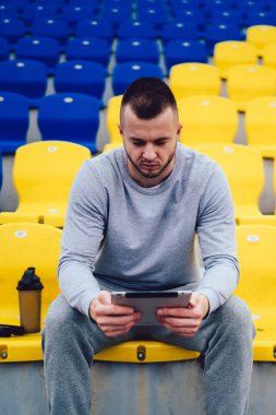 Aktif giyimli genç beyaz erkek sosyal medyadan gelen haberleri tabletle okuduktan sonra dinleniyor, hipster çocuk milenyum koşucusu modern touchpad uygulaması ile sonuçlarını kaydediyor.