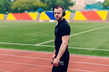 Moda kıyafetleri içinde yakışıklı beyaz bir erkek stadyumda yürüyor favori şarkıları dinliyor, hipster bir adamın portresi ses aksesuarlarıyla online radyodan keyif alarak kardiyo çalışmasına hazırlanıyor.