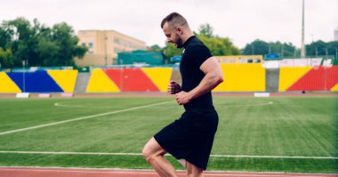 Modern yakın giyimli sporcunun, bulutlu bir yaz gününde stadyumda yüksek dizli koşu yaparken yan görüntüsü.