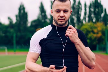 Forma girmiş genç kaslı erkek atlet akıllı telefondan müzik dinliyor bulutlu bir yaz gününde stadyumda koşan kameraya bakıyor.
