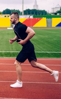Kulaklıklı, aktif giyinmiş, stadyumda koşan genç, yakışıklı bir erkek spor hedeflerine ulaşıyor. Güçlü bir sporcu, sabah kardiyo antrenmanına hazır ve kulaklıklı favori müzikle çıkıyor.