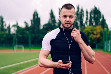 Forma girmiş genç kaslı erkek atlet akıllı telefondan müzik dinliyor bulutlu bir yaz gününde stadyumda koşan kameraya bakıyor.