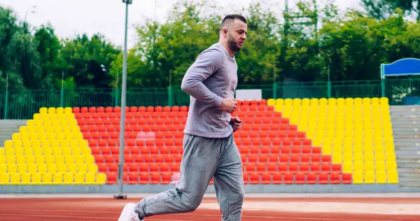 Aktif koşan genç yakışıklı erkek koşucu formda kalmak için sabah antrenmanında stadyumda koşuyor, eşofmanlı kararlı güçlü bir adam dışarıda yağ ve kalori yakmak için antrenman yapıyor.