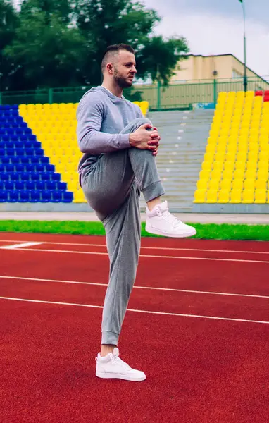 Genç güçlü erkek sporcu ısınmak için egzersiz yapıyor sabah stadyumda ayakta antrenman yaparken gri eşofmanlı 20 'li yaşlarda beyaz sporcu spor yaparken spor yapma hedeflerine ulaşıyor.