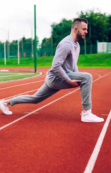 Beyaz tenli erkek koşucunun daha iyi sonuçlar almak ve esnek vücut eğitimi almak için kaslarını esnetirken yan görünüşü. Aktif giyimli yakışıklı genç sporcu stadyumda sabah antrenmanı yapıyor.