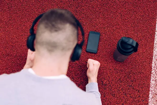 Telefon ve su termosuyla stadyum pistinde antreman yaparken kulaklıkla şarkı dinleyen isimsiz bir gencin yukarıdan görünüşü.