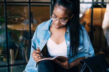 Modayı takip eden siyahi bayanın yukarıdan günlüğe yazarken modern kafenin penceresinin yanındaki göz kamaştırıcı bankta dinlendiğini düşünün.