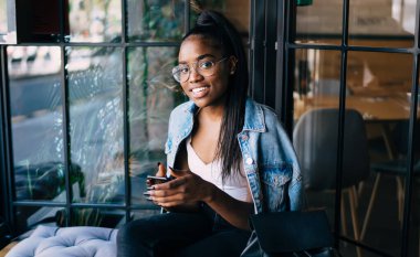Gözlüklü, mutlu ve modaya uygun giyinmiş siyahi bayanın kameraya gülümserken bankta oturması ve cep telefonuyla sörf yapması.