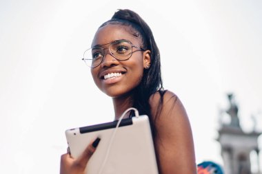 Güneşli bir günde kasabayı keşfetmek için dijital tabletle yürüyen, dokunmatik ped tutan ve gülümseyen, 20 'li yaşlarda Afro-Amerikan genç bir kızın yarım boy portresi.