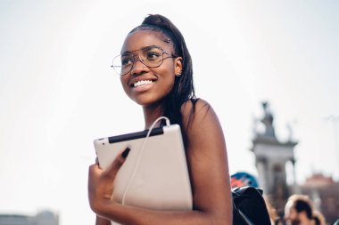 Güneşli bir günde kasabayı keşfetmek için dijital tabletle yürüyen, dokunmatik ped tutan ve gülümseyen, 20 'li yaşlarda Afro-Amerikan genç bir kızın yarım boy portresi.