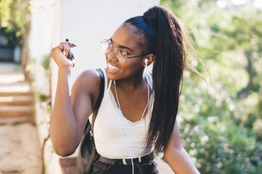 Uzun saçlı, yaz kıyafetleri ve gözlüklü bir kadın güneşli bir günde parktaki korkulukta müzik dinlerken gözlük takıyor ve başka tarafa bakıyordu.