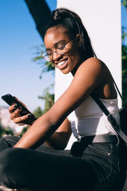 Sırt çantalı, sosyal medyada akıllı telefondan sohbet eden ve gündüz vakti balkonda gülümseyen neşeli Afro-Amerikan kadın.