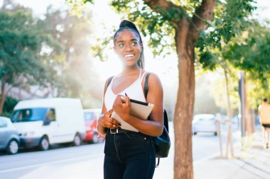 Sırt çantalı, at kuyruklu, tabletli, gün ışığında kaldırımda dikilen siyah kadın.