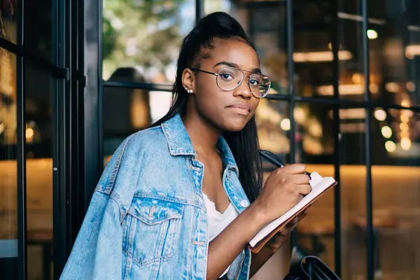 Güzel Afro-Amerikan hippi kız gözlüklü portresi elinde not defteri ve kafenin içinde boş zaman üzerine yazı yazan modaya uygun giyinmiş genç bir kadın kameraya bakıyor ve boş zamanlardaki hobi üzerine çizimler yapıyor.