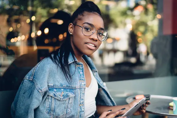4G internete bağlı modern dijital tablet kullanarak kameraya bakan gözlüklü, 20 'li yaşlarda gülümseyen Afro-Amerikan kadın kameralara bakıyor.