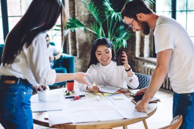 Beyaz tişörtlü neşeli çok ırklı iş arkadaşları iş detaylarını anlatırken ofiste kahve içmek ve kağıtları analiz ederken...