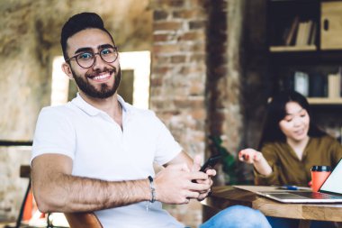 Beyaz tişörtlü ve kot pantolonlu genç adam bilgisayarla aynı masada oturup kameraya bakarken akıllı telefon kullanıyor.