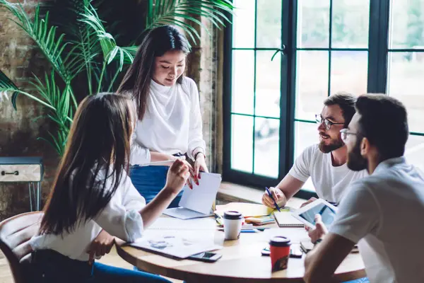 Yaratıcı iş yerinde duran ve iş stratejisini ahşap masada oturan meslektaşlarına açıklayan, günlük kıyafetli Asyalı kadın yönetici.