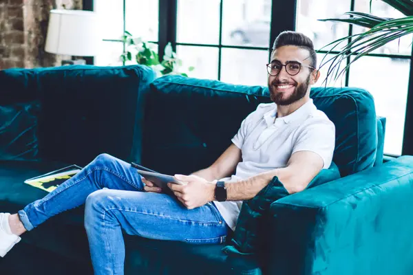 Uzun boylu, mutlu, gözlüklü, rahat koltukta oturan ve loft ofisindeki tableti kullanan etnik bir adamın yan görüntüsü.