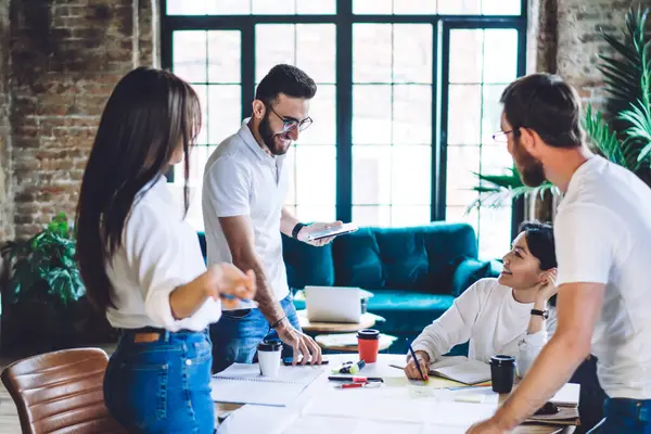 Neşeli çok ırklı meslektaşlar, ahşap masa etrafında kağıtlar ve hazır fincanlarla toplanarak modern çalışma alanında tuğla duvarlı projeleri tartıştılar.