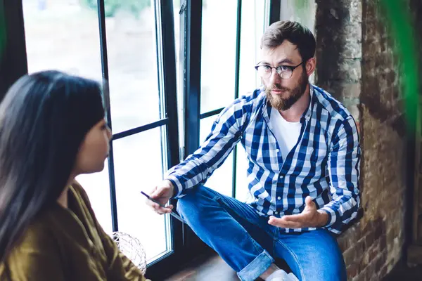 Üstünde günlük giysiler ve gözlüklerle pencerenin kenarında oturan ve ofisteki iş projesiyle ilgili konuşan konsantre bir adamdan.