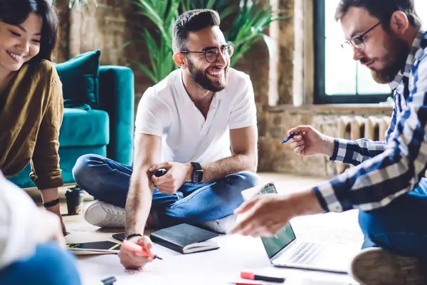 Yaratıcı çalışma alanında çalışırken ve yerde otururken gülümserken gündelik kıyafet çizimi projesinde çalışan çok ırklı meslektaşlar.