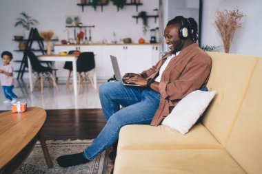 Kulaklıkla gülümseyen, koyu tenli, kulaklık takan, ev içi internet sitesi için laptop teknolojisi kullanan, elektronik cihazlı mutlu Afrikalı Amerikalı adam, netbook 'ta müzik podcast' i arıyor.